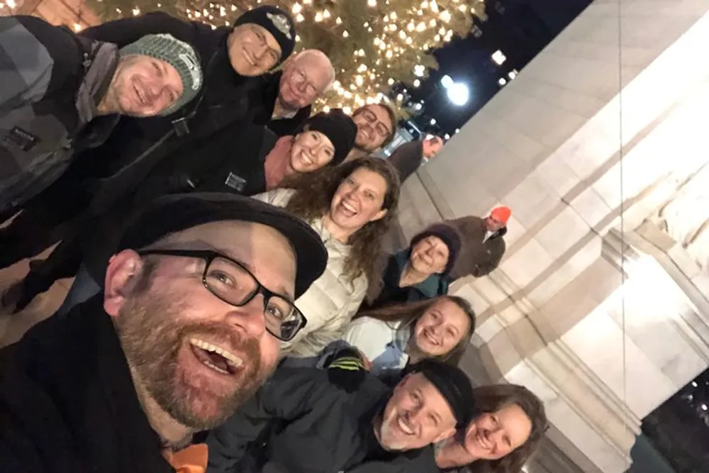 A group of smiling people is taking a cheerful group selfie at night with twinkling lights in the background