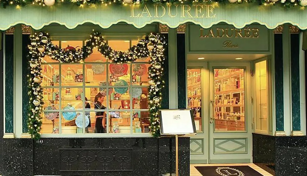 The image shows the elegant festively decorated exterior of a Ladure patisserie renowned for its macarons with a glimpse of the interior and an employee visible through the window