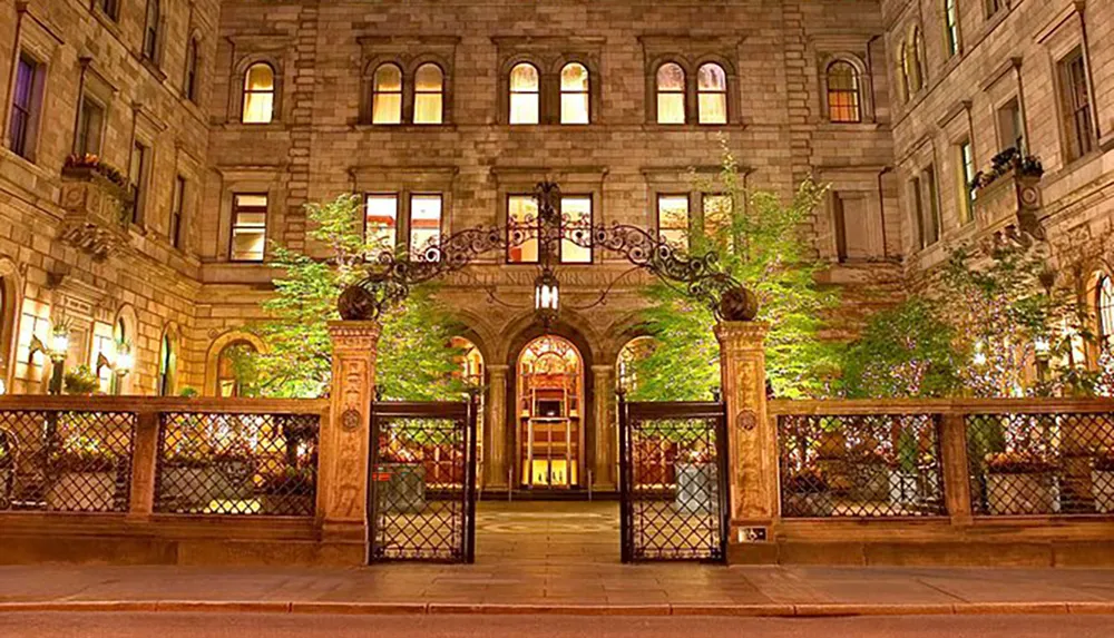 The image features a warmly lit entrance of a classic stone building with ornate iron gates and a balcony adorned with foliage during the evening