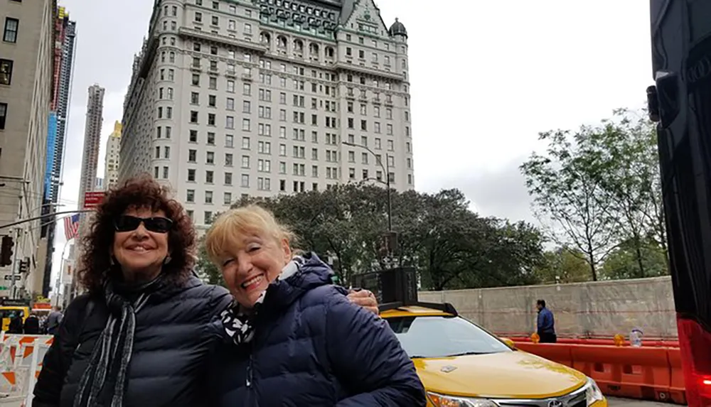 Two smiling women pose for a photo on a city street with a distinctive historic building in the background and a taxi in the foreground