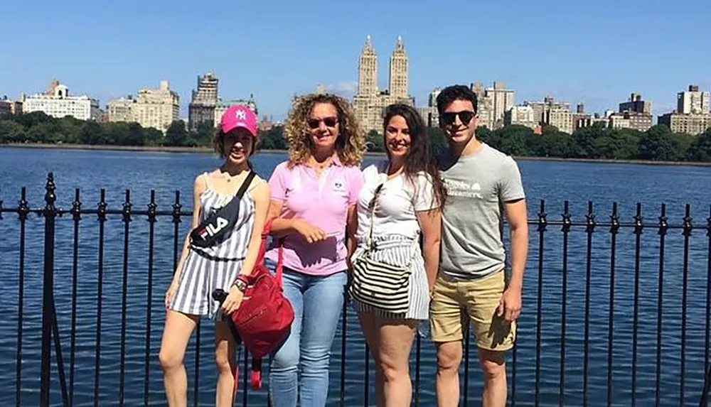 Four people are smiling for a photo in front of a lake with a city skyline in the background on a sunny day