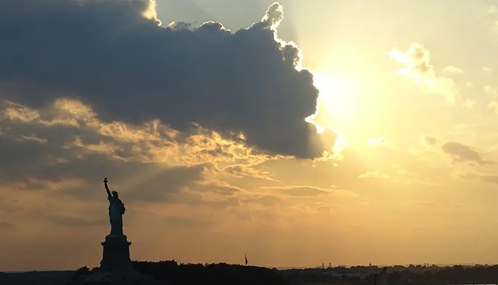 The silhouette of the Statue of Liberty is set against a dramatic sunset with the sun peeking out from behind cloud cover