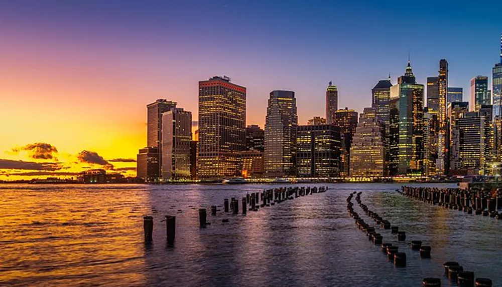 The image captures a stunning sunset behind the skyscrapers of a city skyline with glowing reflections on the water and old pier structures leading into the river