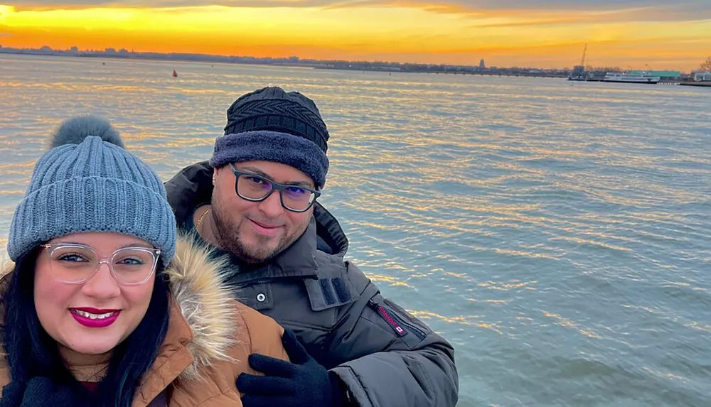 Two smiling people wearing winter hats and jackets are posing for a selfie with a waterfront view at sunset