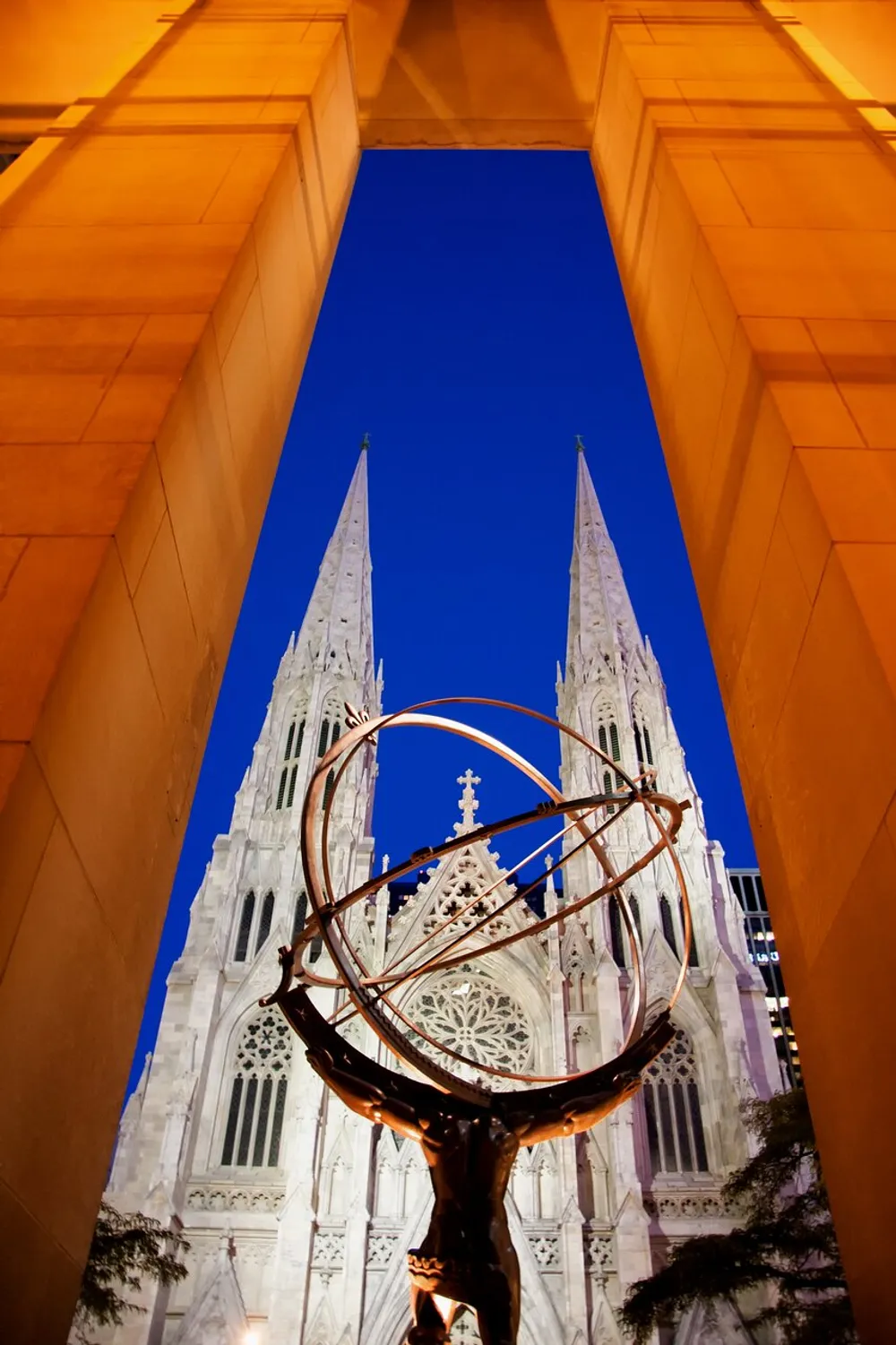 The image shows the towering spires of a Gothic cathedral framed by a dramatic archway and contrasted with a modern sculpture in the foreground against a deep blue sky