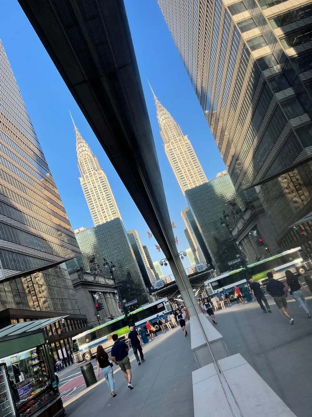 The image captures a bustling city street with pedestrians showcasing a reflective building surface that creates a mirrored illusion of the distinctive Chrysler Building against a clear blue sky