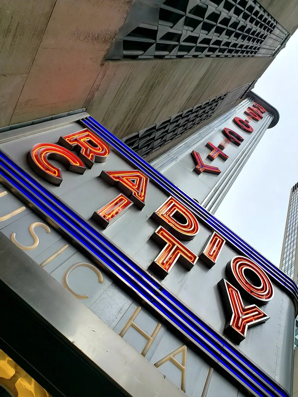 The image shows a low-angle view of the Radio City Music Hall marquee with the iconic neon sign set against the backdrop of a building in an urban environment