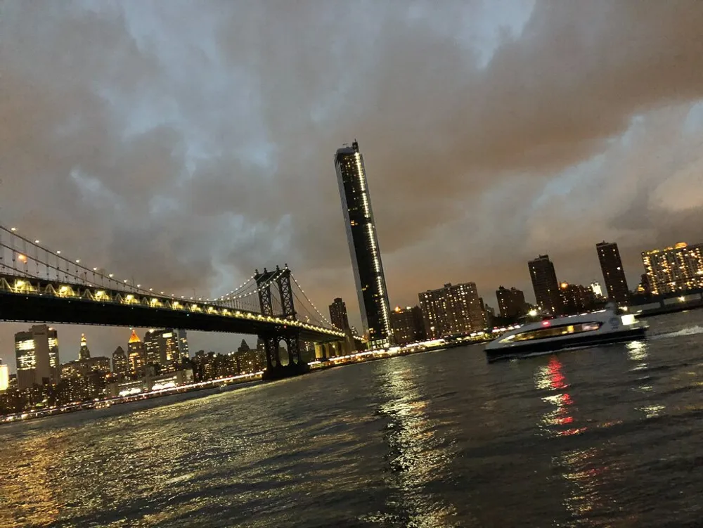 An evening view of a lit-up suspension bridge with a city skyline in the background under a cloudy sky as seen from the waterfront with a speeding boat passing by