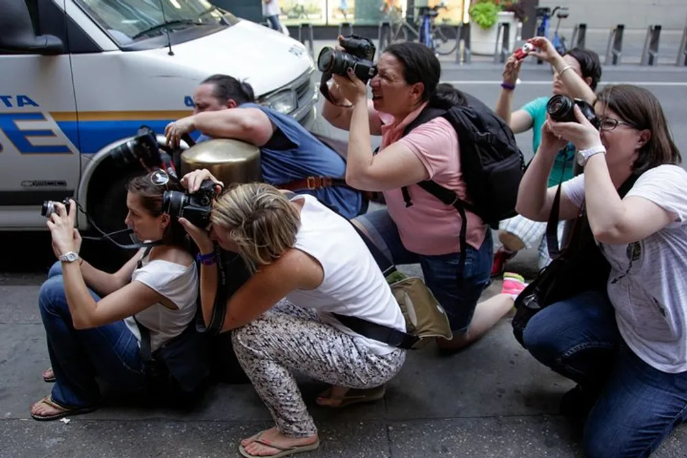 A group of photographers is crouched down aiming their cameras towards something of interest out of the frame near a police vehicle on the street