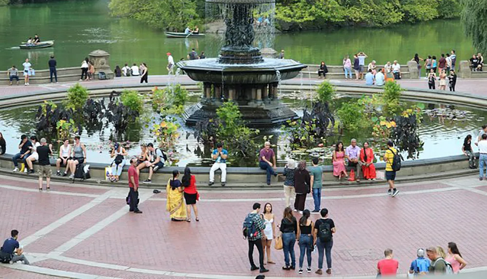 Visitors are enjoying a sunny day around a large fountain with a pond in the background where people are boating