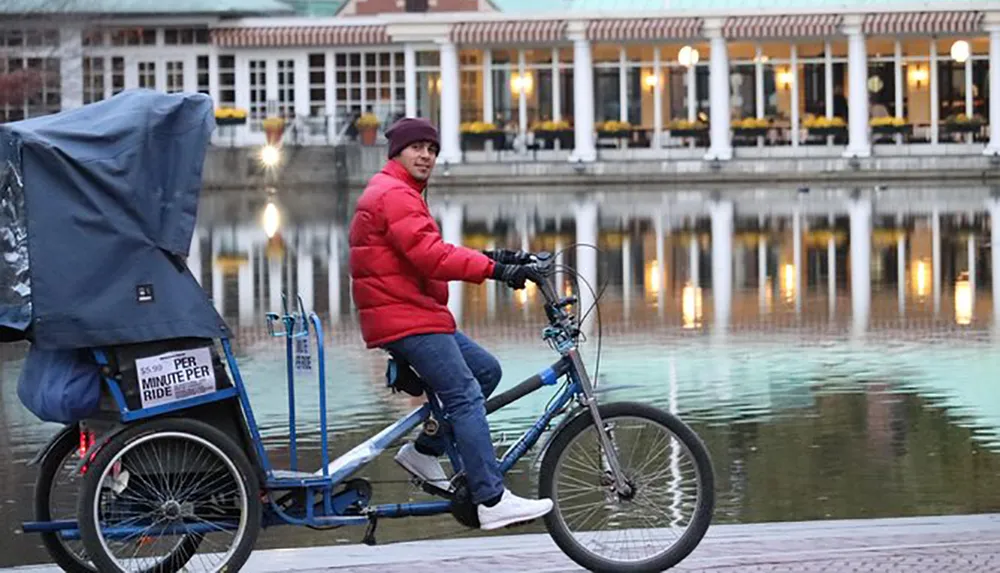 A pedicab driver in a red jacket is looking back at the camera while riding his bike along a waterside promenade with lit buildings reflected in the water