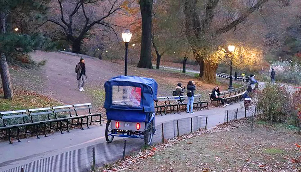 A pedicab is parked beside an illuminated pathway in a park where people are walking and sitting on benches during what appears to be evening or twilight
