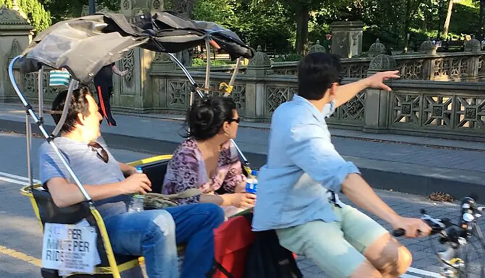 Three people are seated on a makeshift bike-powered vehicle with an umbrella on a sunny day while the person in the front gestures towards something out of frame