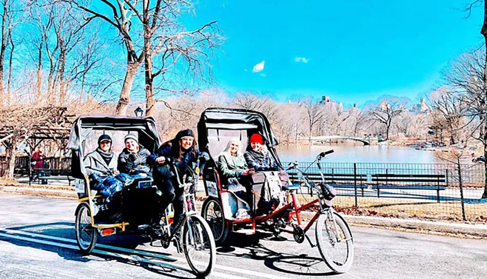 Two pedicabs carrying passengers ride along a park path near a lake on a sunny day