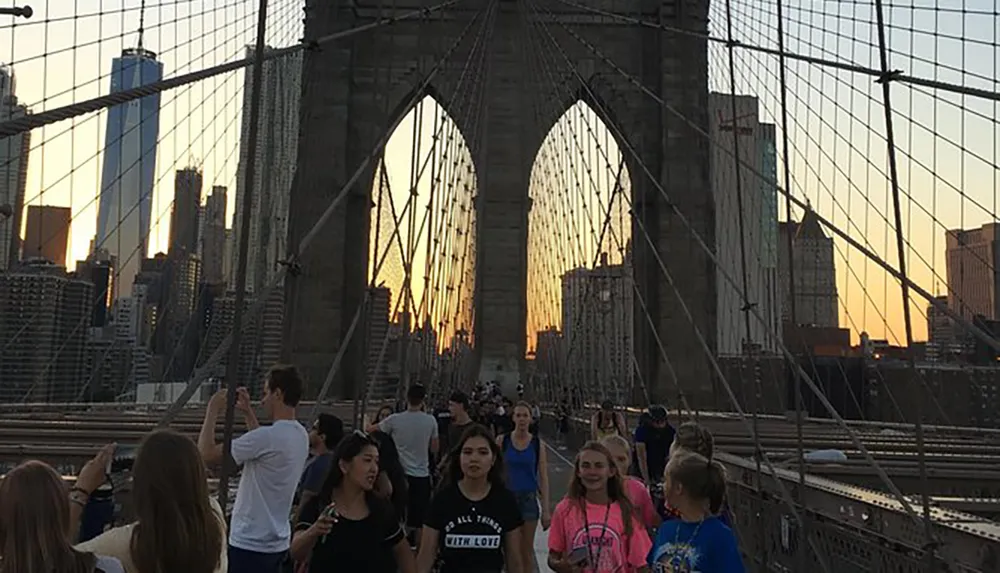 Pedestrians are enjoying a walk on the Brooklyn Bridge at sunset with the Manhattan skyline in the background