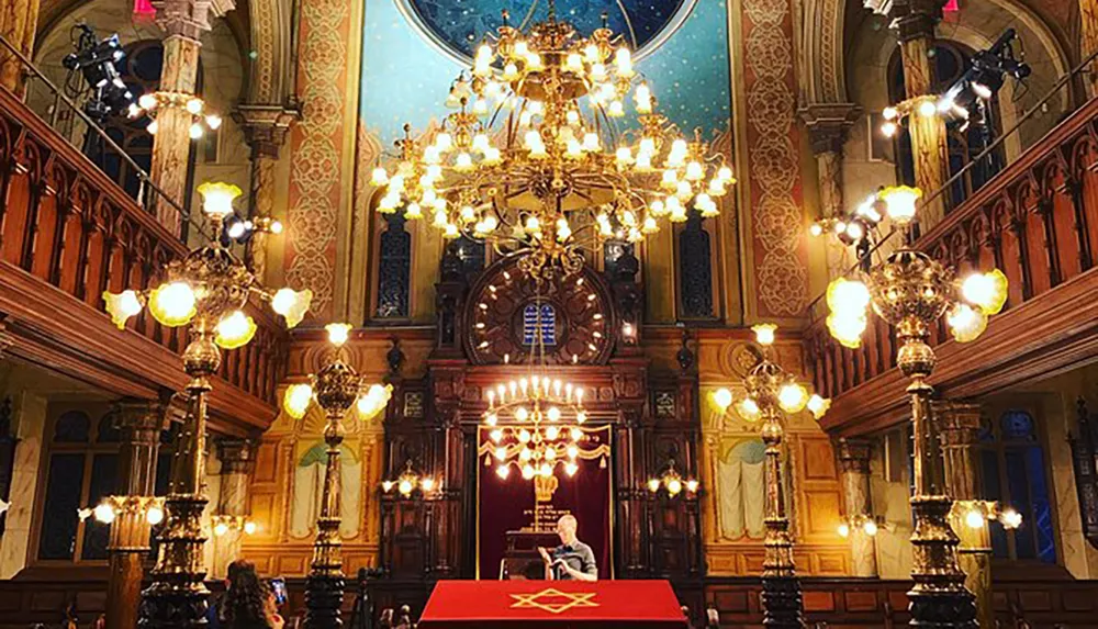 The image shows an ornately decorated synagogue interior with a person seated at a podium beneath an elaborate chandelier surrounded by traditional Jewish symbols and intricate architectural details