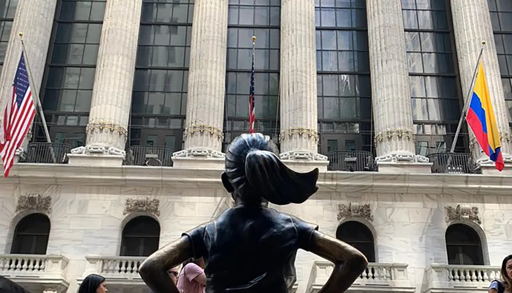 The image shows the Fearless Girl statue facing the New York Stock Exchange building with flags including the American and Colombian flags flying overhead