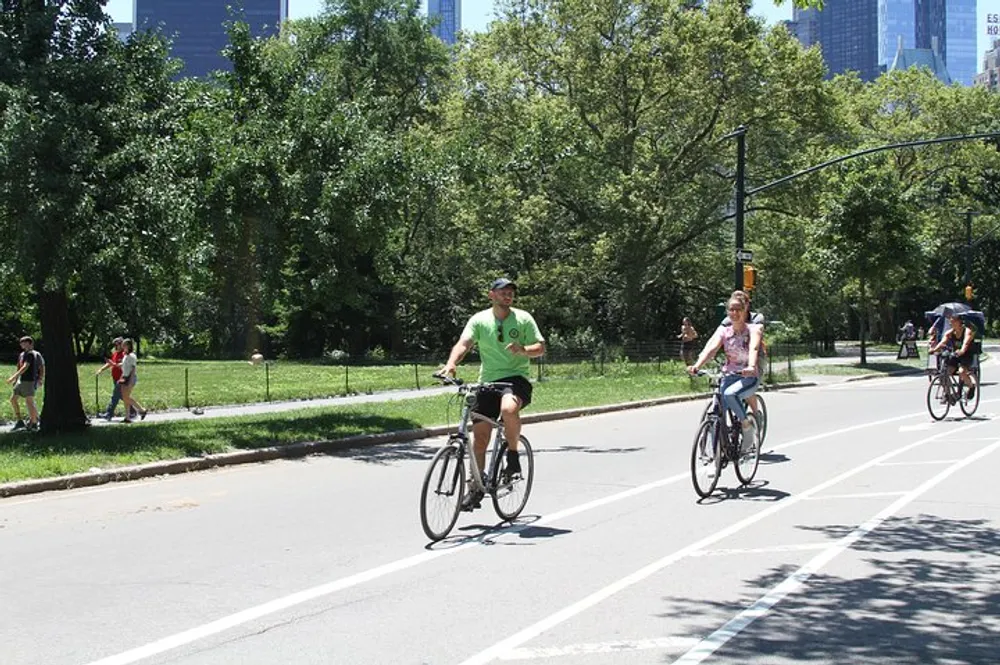 People are enjoying a sunny day bicycling in a park with trees and a clear blue sky with a glimpse of tall buildings in the background