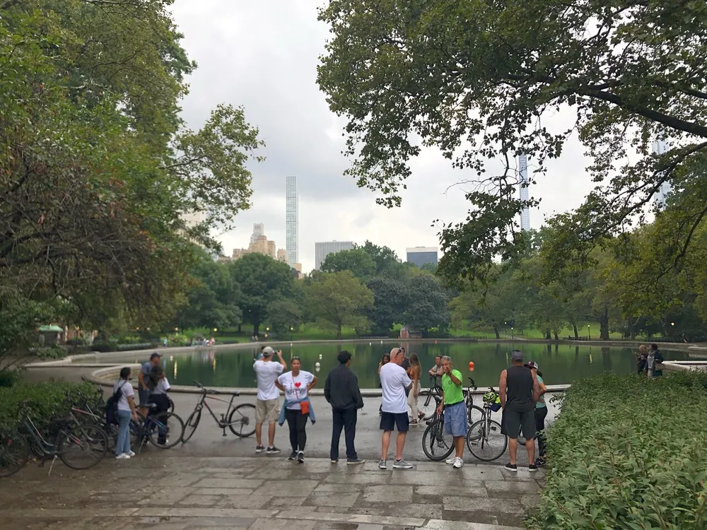 Several people including cyclists are gathered around the edge of a tranquil pond in a leafy park with skyscrapers towering in the background