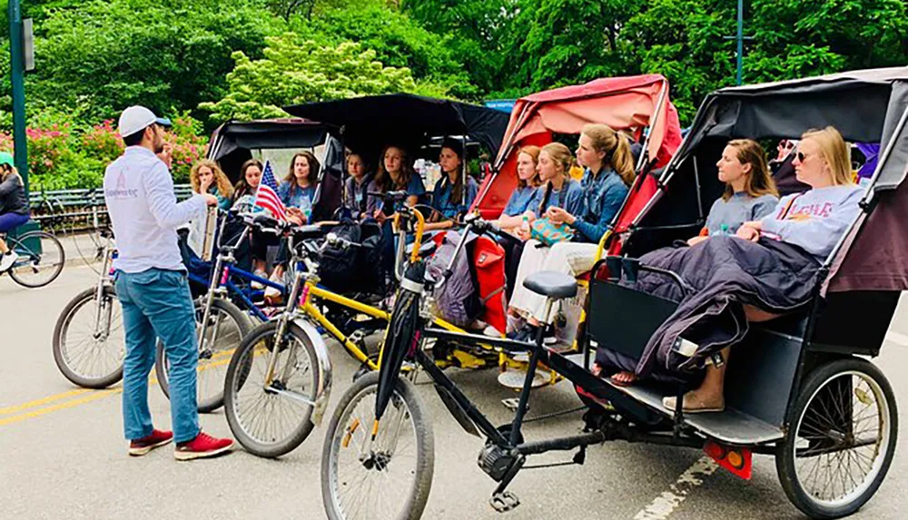 A group of people are sitting in pedal-powered rickshaws listening to a guide who is standing and addressing them