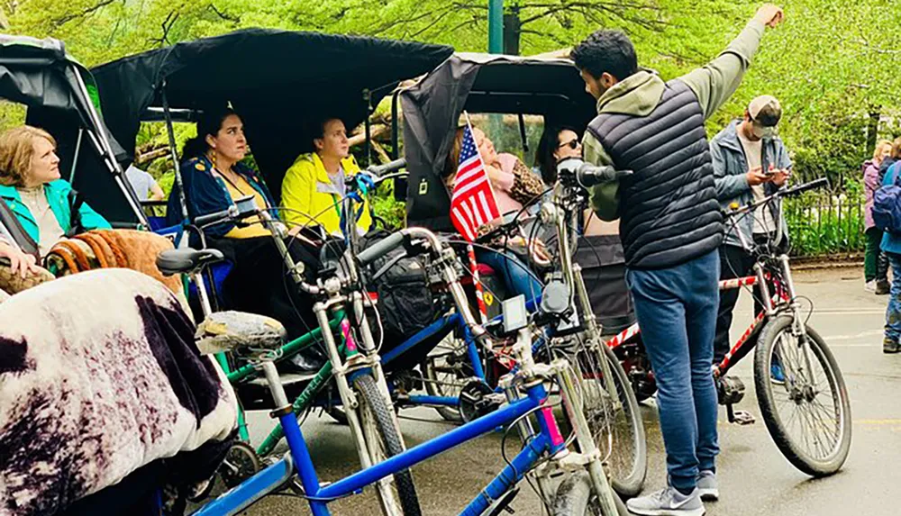 This image shows a man pointing something out to seated passengers in pedicabs with various people and bicycles visible suggesting a tour or ride service in an urban park setting