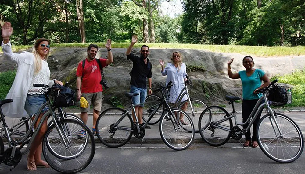 A group of people are smiling and waving to the camera while standing next to their bicycles in a park setting