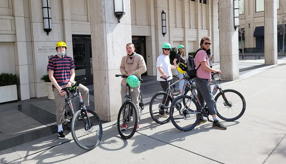 Five individuals wearing helmets are standing with their bicycles on a sunny day in an urban setting
