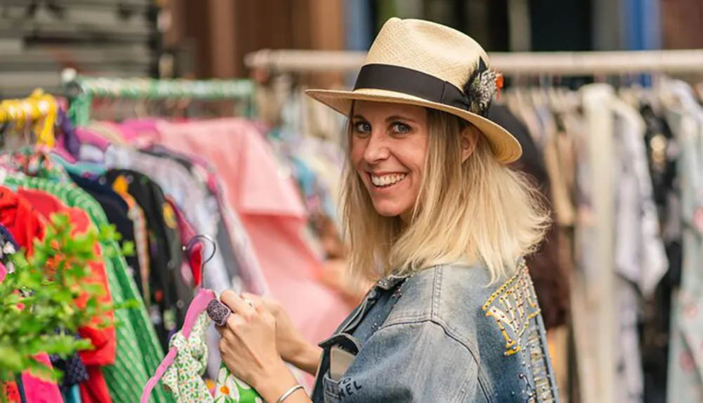 A smiling woman wearing a hat is browsing through clothing at an outdoor market