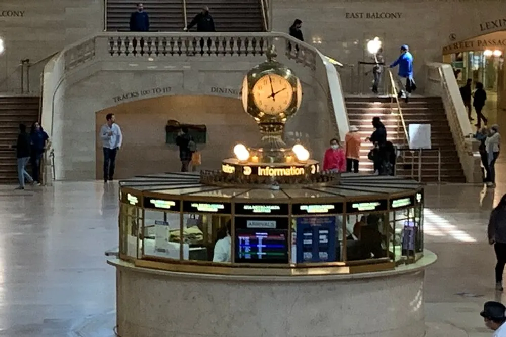 The image depicts the iconic information booth with a golden clock at the center of Grand Central Terminal in New York City with people milling about in the background