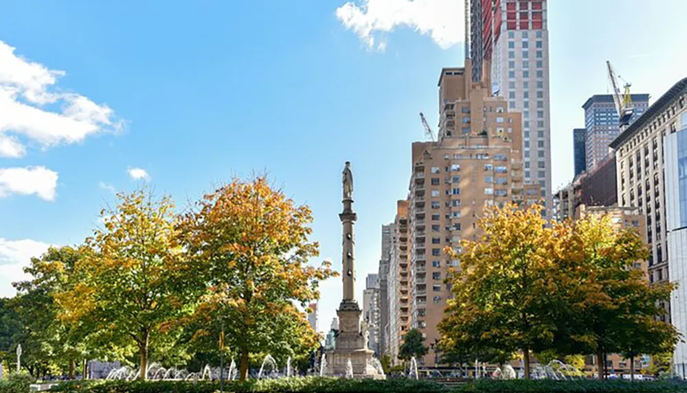 The image shows a cityscape with autumn-colored trees in the foreground a monumental column with a statue on top in the middle and a backdrop of mixed architecture under a blue sky with scattered clouds