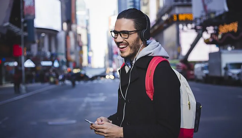 A happy man wearing headphones and glasses is holding a smartphone on a bustling city street
