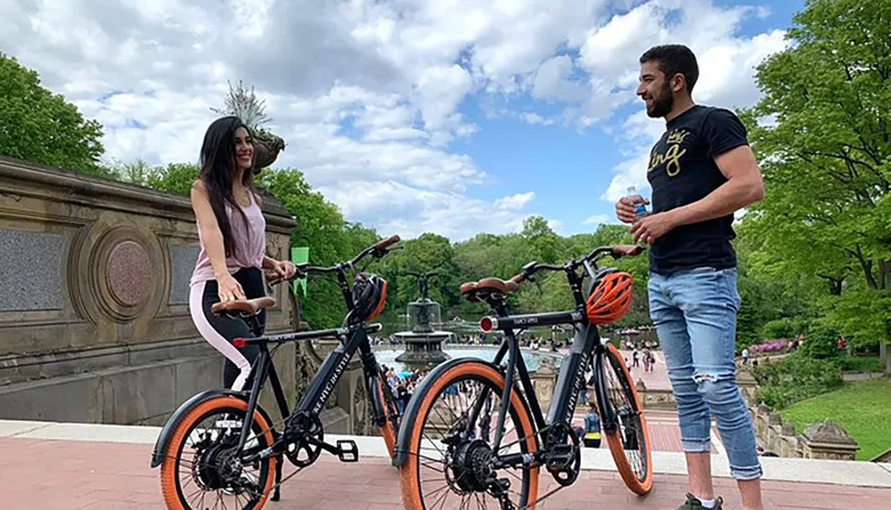 Two people stand next to their bicycles in a park smiling and holding water bottles with a fountain and trees in the background