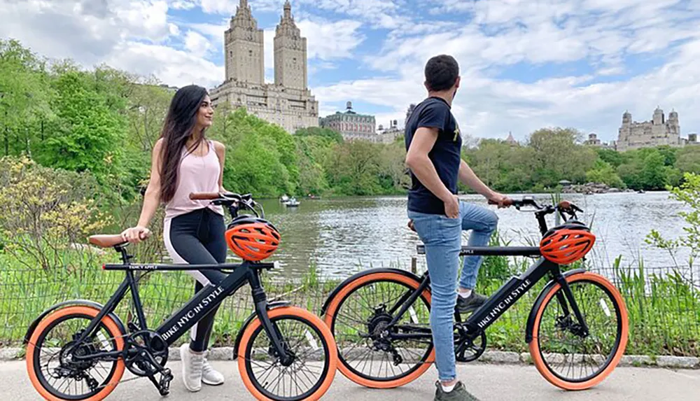 A man and a woman stand next to their bicycles by a lakeside with urban buildings in the background