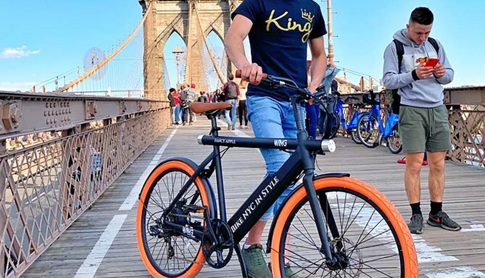 A person rides a stylish bicycle with orange wheels on a busy bridge while another person stands looking at their phone