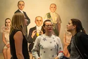 Three women are engaged in a conversation in an art gallery with surreal paintings of figures with oversized heads in the background.