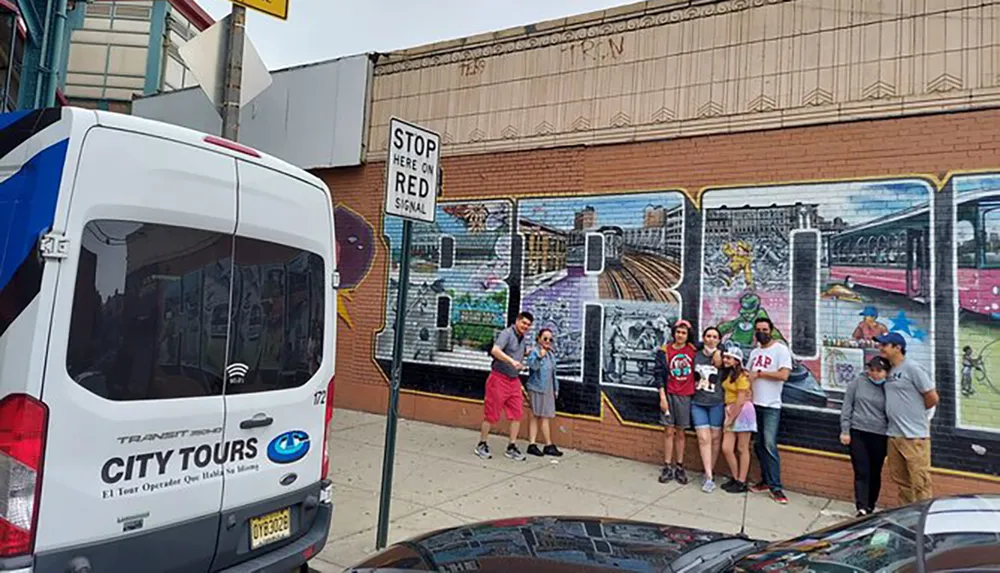 A group of people poses for a photo in front of a colorful urban mural while a city tour van is parked nearby