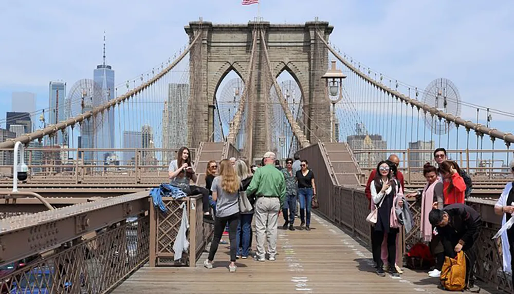 The image shows pedestrians walking and taking photos on the Brooklyn Bridge with the New York City skyline in the background