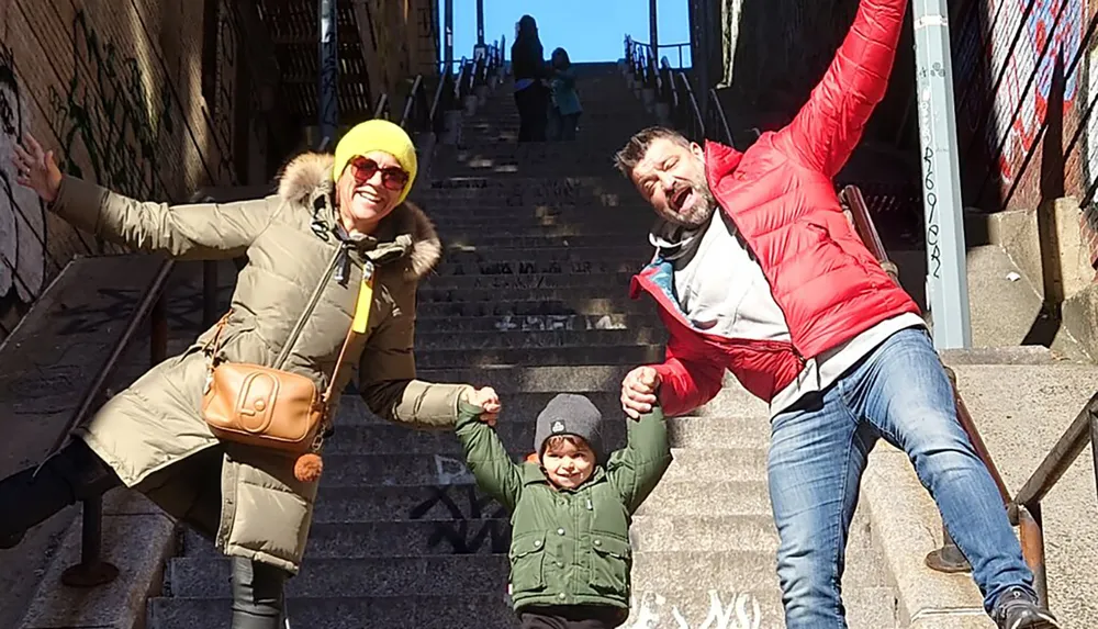 A family of three is joyfully posing on a set of urban stairs with two adults holding hands with a child all smiling brightly in the sunlight
