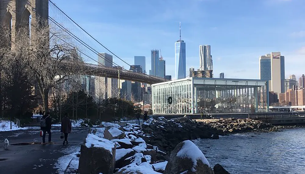 The image captures a snowy riverfront scene with pedestrians a dog the Brooklyn Bridge and the Manhattan skyline in the background