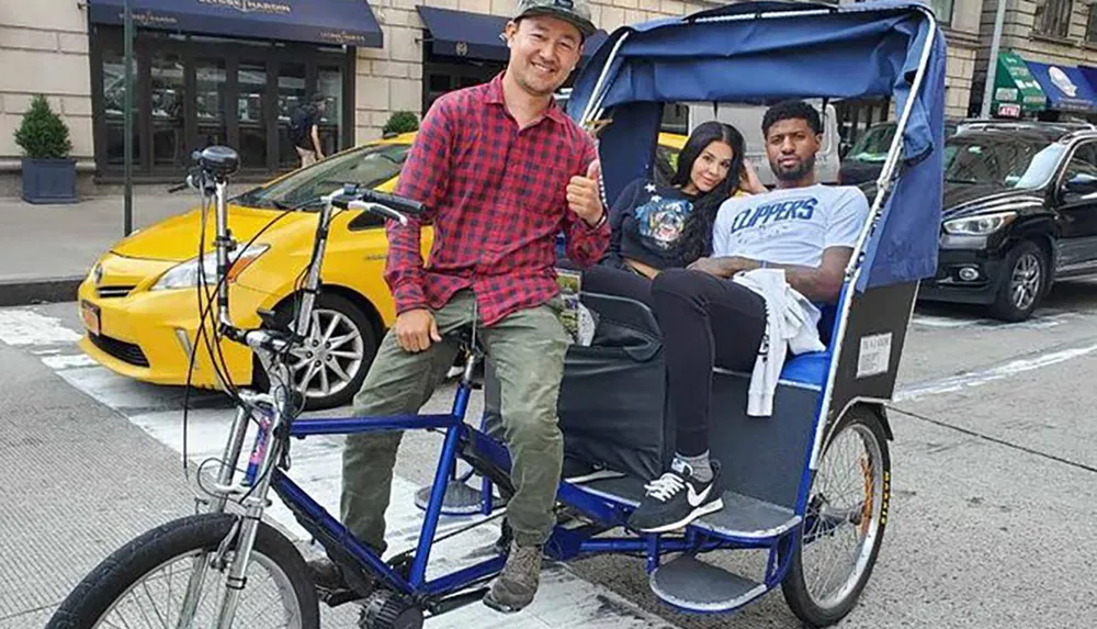 A pedicab driver is giving a thumbs-up next to his vehicle while a man and a woman sit in the passenger seat on an urban street