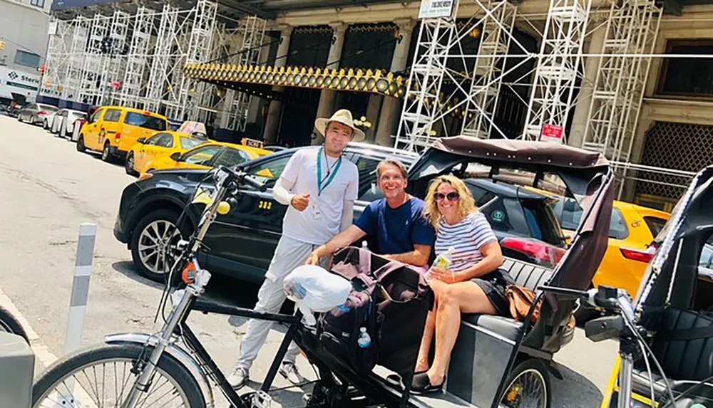 Two tourists are posing for a photo in a pedicab with their driver surrounded by yellow taxis on a sunny day in an urban setting