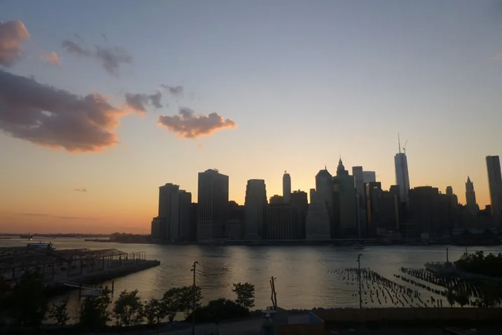 The image captures a tranquil sunset behind the silhouette of a city skyline possibly New York with reflections on the water and scattered clouds in the sky