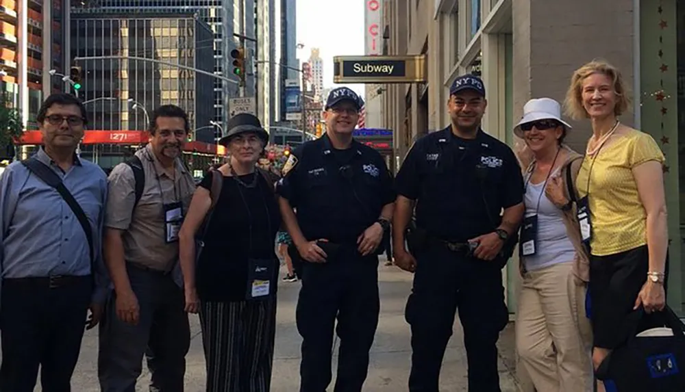A group of tourists pose for a picture with two NYPD officers on a city street with a subway entrance in the background