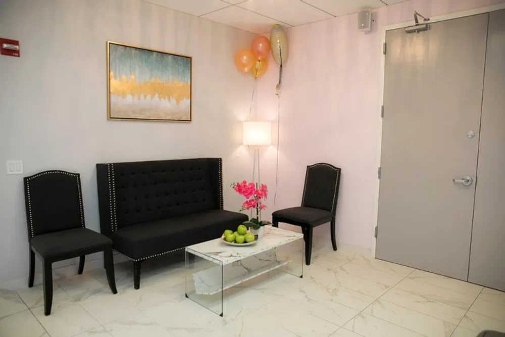 A modern waiting room with a black couch and chairs a white marble table with fruit on it pink flowers balloons and a painting on the wall