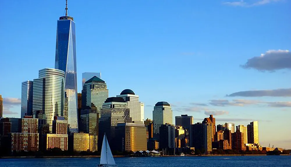 The image captures a view of the Lower Manhattan skyline featuring the One World Trade Center with a sailboat floating in the foreground under a blue sky