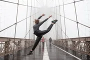 A person is performing a high kick on a foggy bridge with pedestrians in the background.