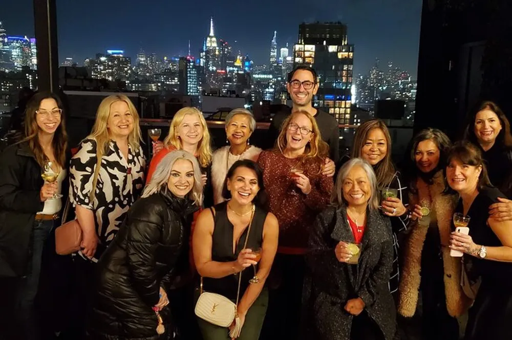 A group of people are smiling and posing for a photo at a night-time rooftop gathering with a city skyline illuminated in the background