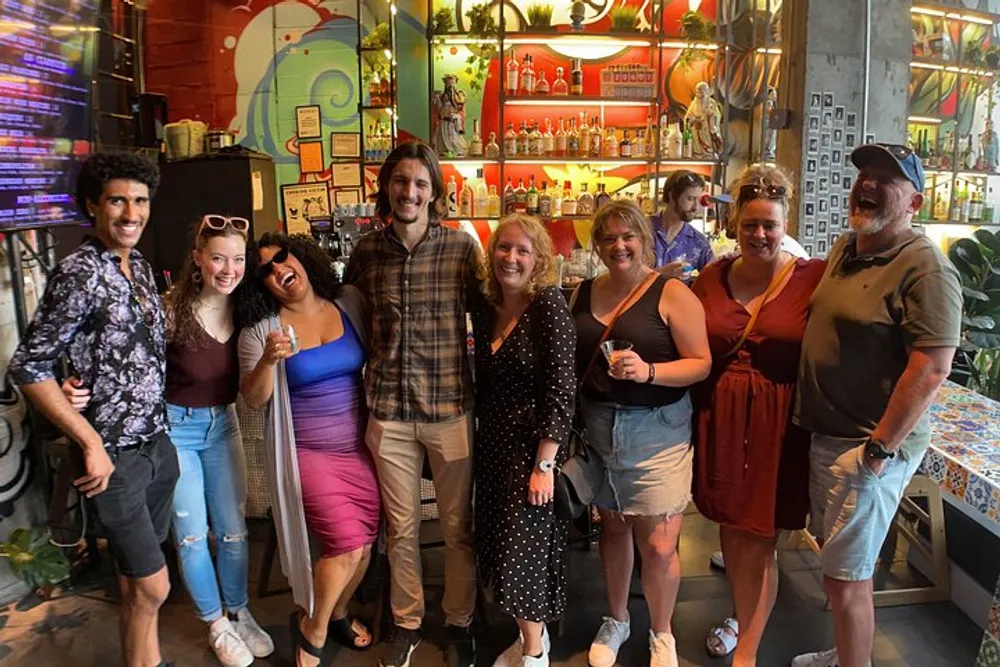 A group of happy people is posing for a photo in a vibrant bar setting with colorful decor and a variety of beverages displayed in the background