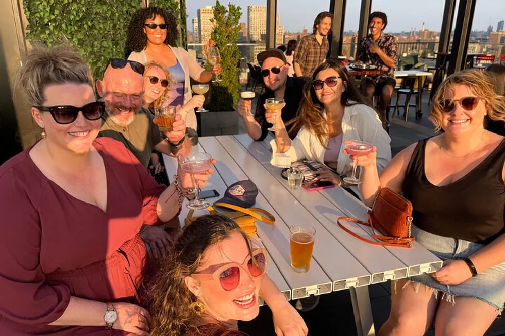A group of people are enjoying drinks and smiling at an outdoor rooftop gathering during sunset