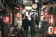 A couple is walking down a vibrant alley illuminated by neon signs and traditional lanterns, evoking the bustling atmosphere of an urban Japanese dining district.